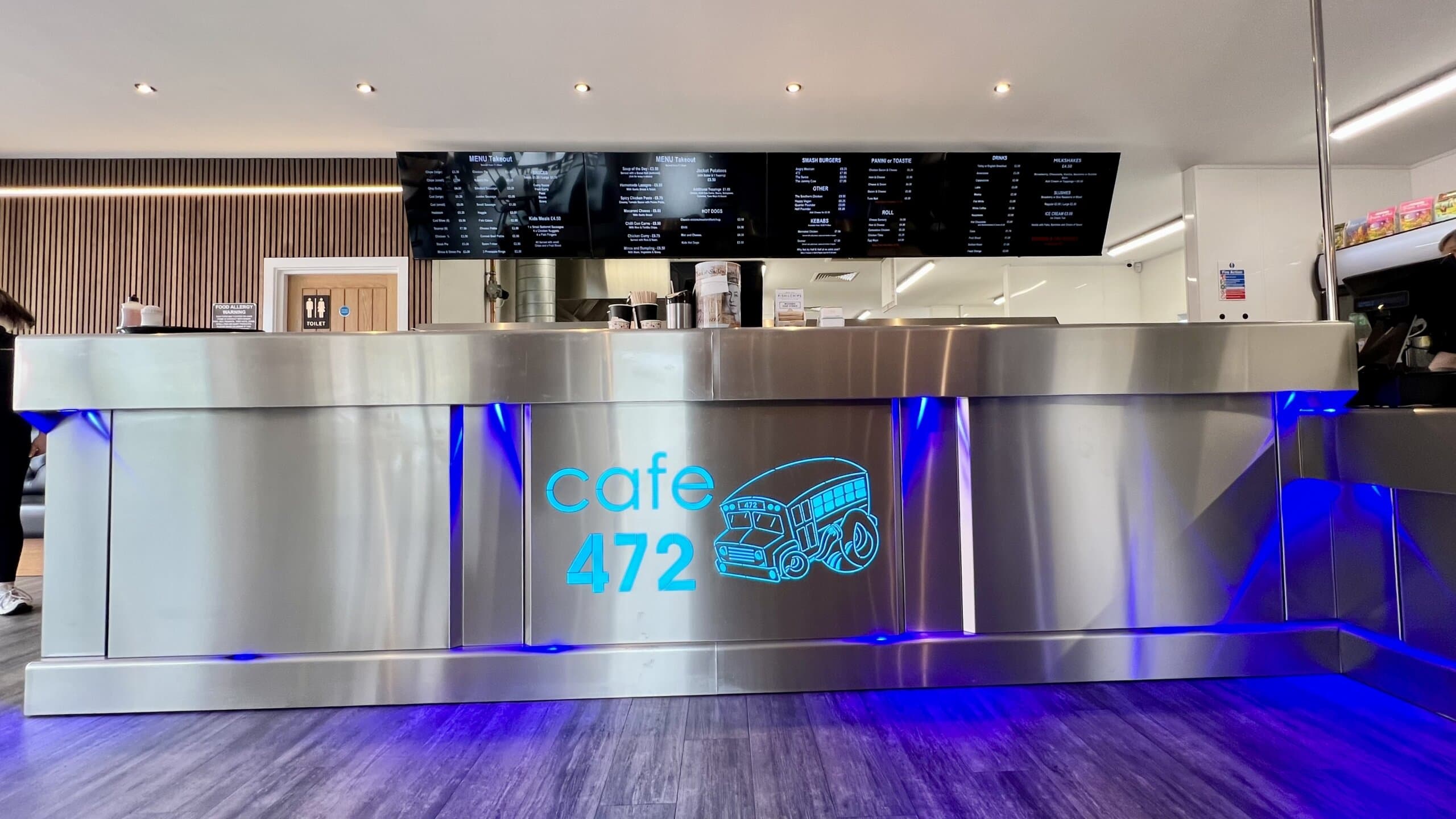 Modern stainless steel café counter with blue LED lighting at Cafe 472, featuring digital menu boards and a contemporary interior design for a trendy dining experience.